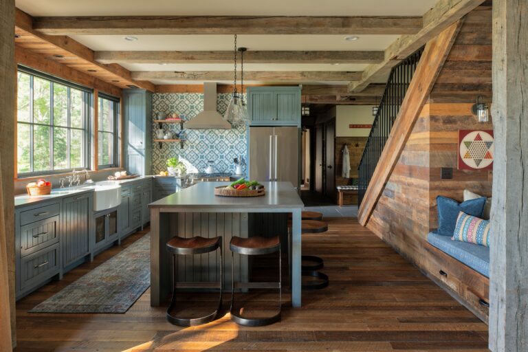 A rustic kitchen with wooden floors, blue cabinets, a patterned backsplash, and reclaimed wood paneling for walls. Large windows, an island with stools, and a cozy built-in bench with pillows under a staircase fill the space with natural light.