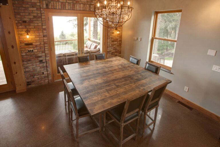 A rustic dining room with a large table with a wood tabletop, eight metal-framed chairs, a brick accent wall, a modern chandelier, and large windows offering views of trees and a balcony with outdoor seating.