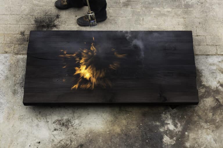 A man holding a fire torch and burning a big piece of oak creating flames and smoke, old jappanese shou sugi ban technique. black wooden material. Grain, texture