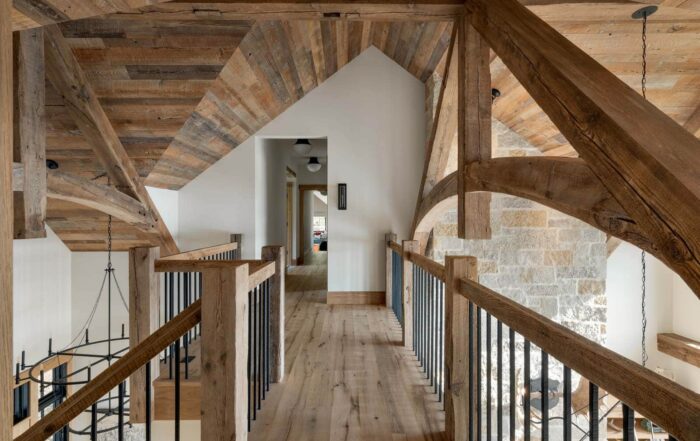 A rustic hallway with exposed wooden beams, wood walls, a wooden walkway with black metal railings, a vaulted wood-paneled ceiling, and stone walls leads to a modern, well-lit room in the distance.