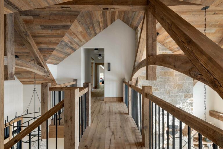 A rustic hallway with exposed wooden beams, wood walls, a wooden walkway with black metal railings, a vaulted wood-paneled ceiling, and stone walls leads to a modern, well-lit room in the distance.