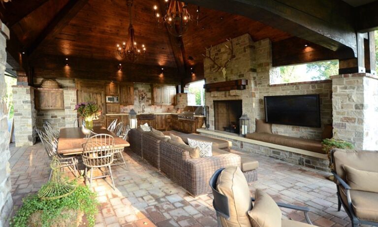 A spacious outdoor living area with a high wooden ceiling, stone walls, wicker sofas, a dining table, fireplace, mounted TV, and full kitchen. Reclaimed wood accents enhance the rustic-meets-modern interior design and provide ample seating.