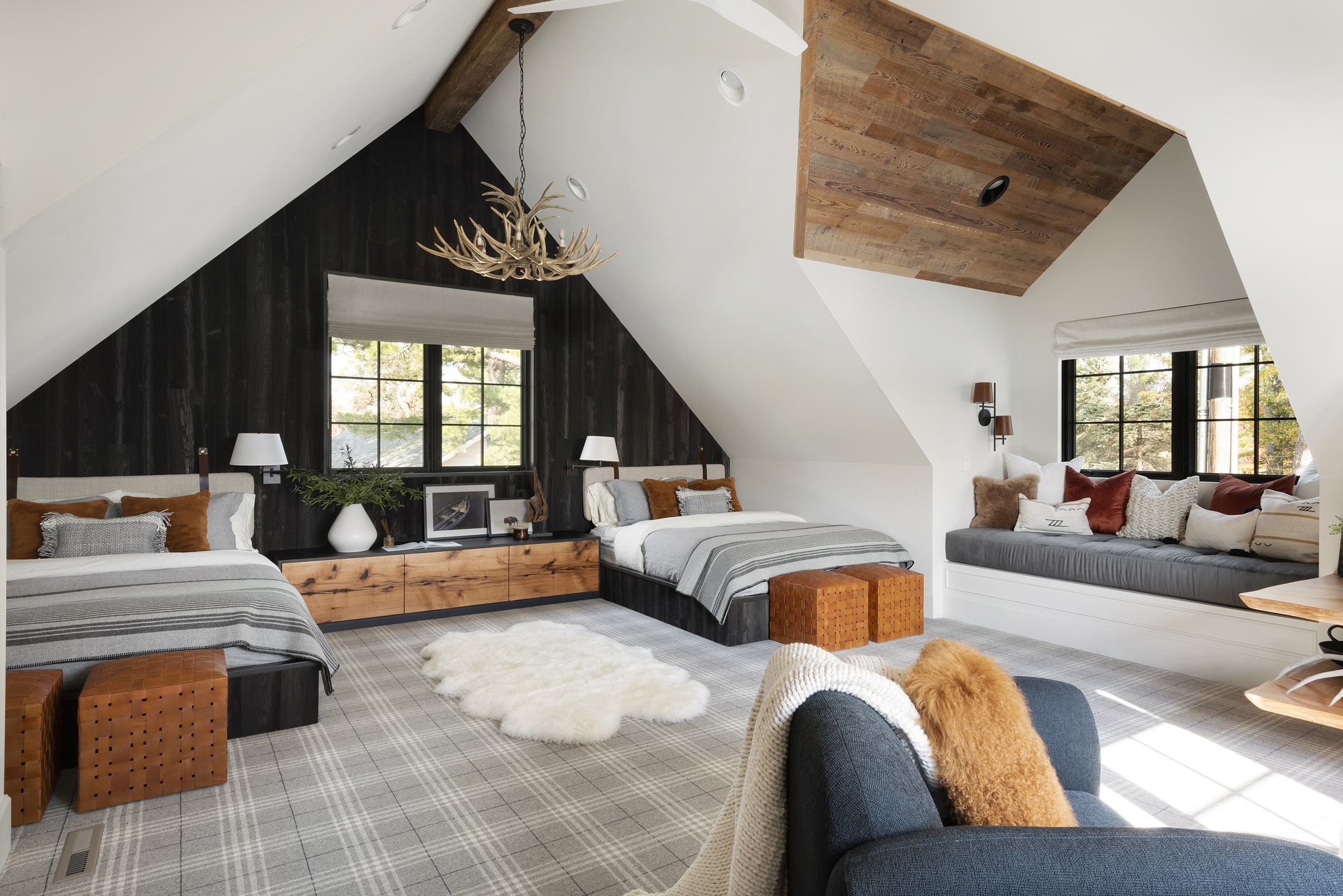 A cozy attic bedroom by Wes Hanson Builders features two beds, a window seat, plaid carpet, rustic wood accents, an antler chandelier, and warm-toned pillows for a modern cabin atmosphere filled with natural light from large windows.