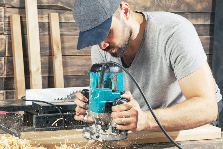 A young man in a gray cap and a gray T-shirt carpenter builder holds a milling machine in the hand and equips the edges of the wooden board in the workshop, wooden sawdust flies to the sides