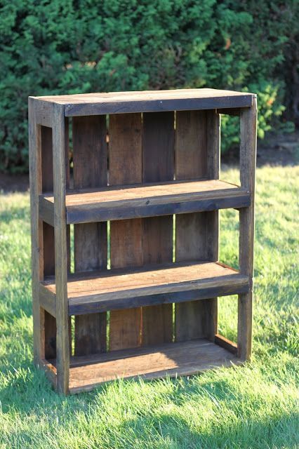 A simple, rustic reclaimed wood bookcase with three shelves stands on grass outdoors, green bushes in the background. The weathered, stained wood gives the shelf a charming vintage look.