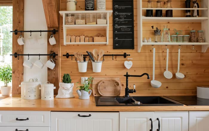 A cozy kitchen with wood cladding, white cabinets, a black faucet, and open shelves holding cups, jars, utensils, and plants. Pots and kitchen tools hang on rods above a wooden countertop and black sink.