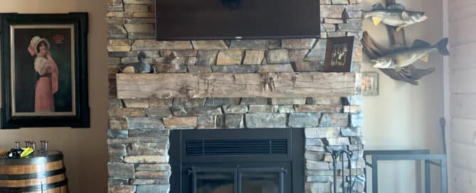 Reclaimed wood mantel above a wood burning fireplace insert. A large TV is mounted on the stone surround above the mantel. Hardwood floors and wood panel ceilings enclose the room.