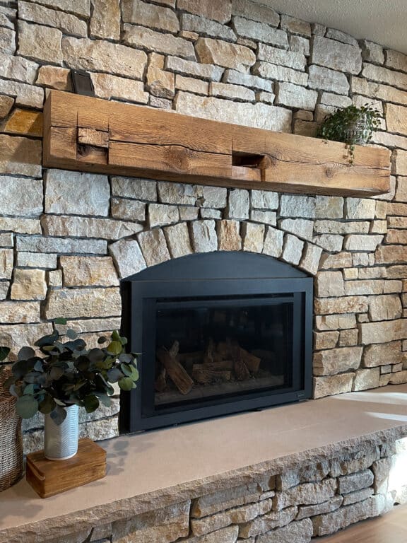 A hand-hewn reclaimed wood mantel over a stone faced fireplace. Decorative potted plants accent the hearth.