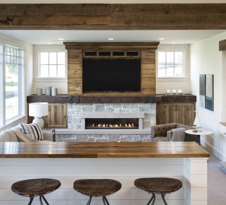 An extended, dark reclaimed wood mantel with iron bands connects two walls. A wide fireplace with a stone surround is below and a TV is mounted to a wood paneled wall above. 