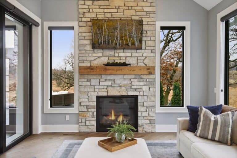 A reclaimed wood mantel against light stone and overhanging a new gas fireplace. Bright natural light shows clean grey and white walls and windows.