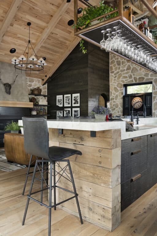 A contemporary bar area made with reclaimed wood and custom reclaimed wood furniture. The bar is made from reclaimed pine timbers and has a marble counter and hanging display shelf above it with stemware racks. In the background more reclaimed wood paneling covers the walls and high ceiling.