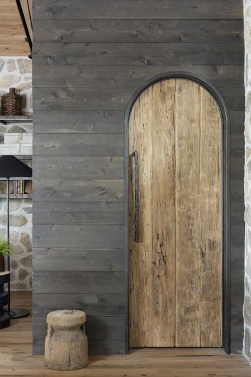 Custom reclaimed wood door faced with hand hewn timbers. The door is narrow with a rounded top and sits against a wall of darker stained wood panels.