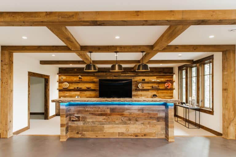 Reclaimed wood bar with reclaimed wood panels above and behind as well as reclaimed wood box beams and pillars accenting the room. A TV is behind the home bar, and blue LED lights illuminate the underside.