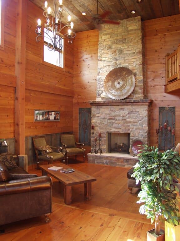 A grand room in a northern lodge with a double high ceiling and large, stone fireplace against one wall. The floors and walls are done in Douglas Fir reclaimed wood paneling.