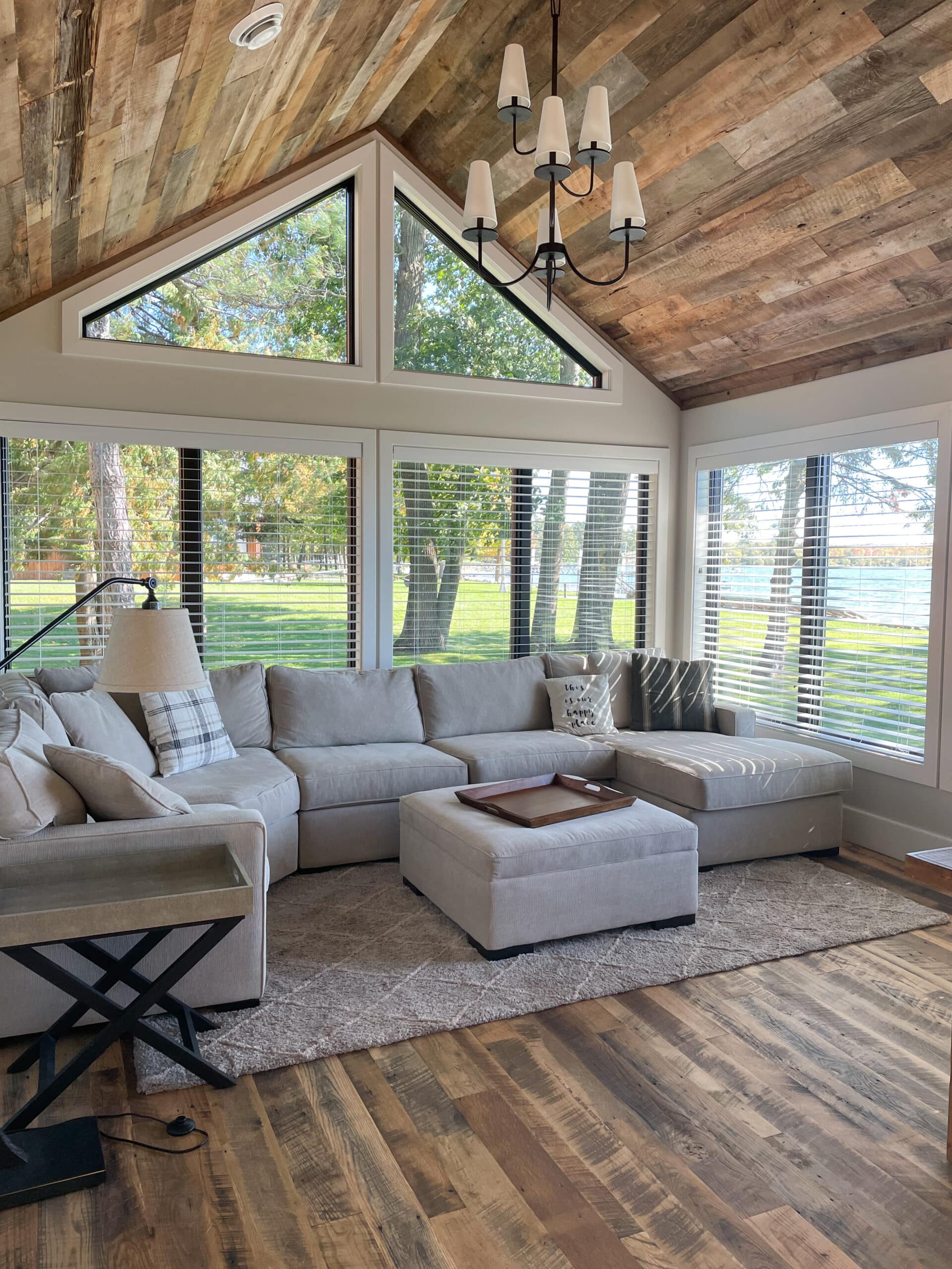 A bright sunroom with a cozy gray sectional and a green yard through the windows. The ceiling is made from reclaimed wood paneling in weathered antique.
