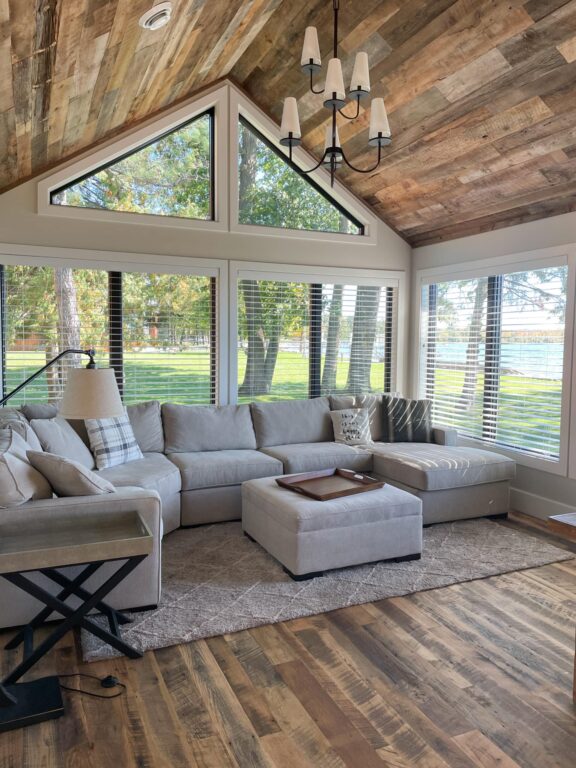 A cozy living room with a large sectional sofa, ottoman, and plaid pillow sits on a rug. Large windows and a wooden ceiling with wood paneling let in natural light and reveal a view of green trees and grass outside.