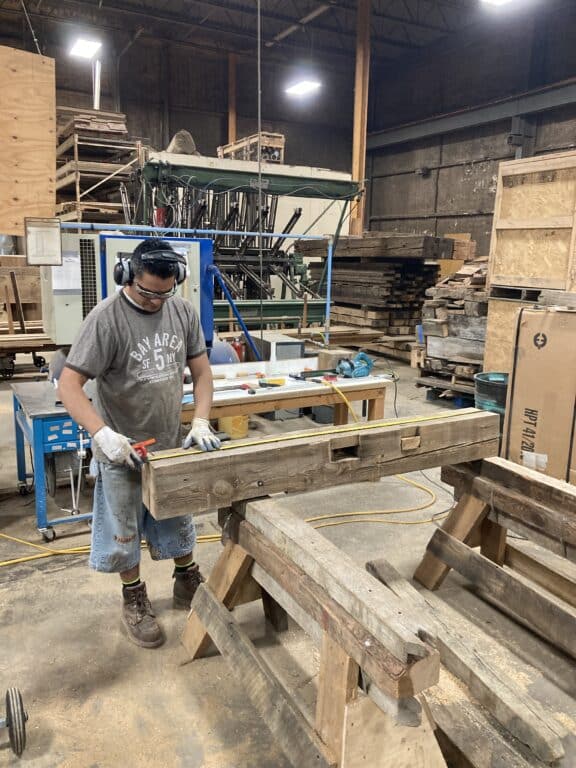 A man wearing safety glasses, gloves, headphones, a t-shirt, and jean shorts works on a piece of reclaimed wood in a workshop filled with tools, machinery, and stacks of lumber.