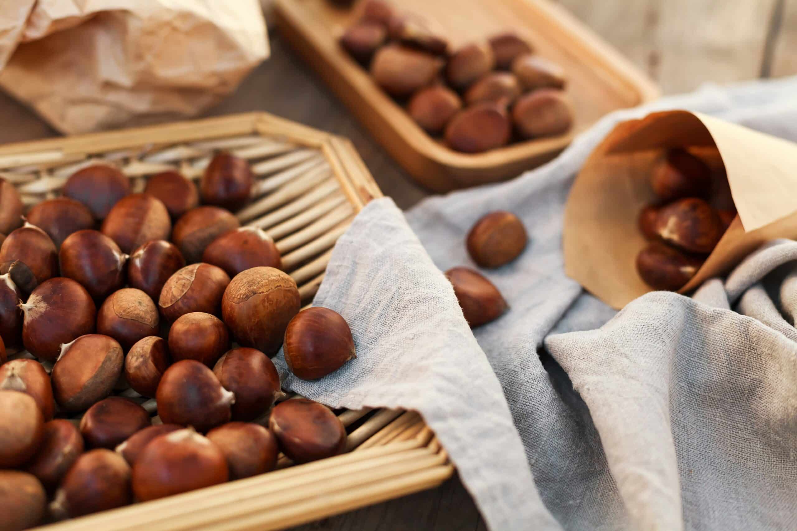 A shallow wicker basket full of chestnuts next to a paper bag with more chestnuts on top of a blue linen.