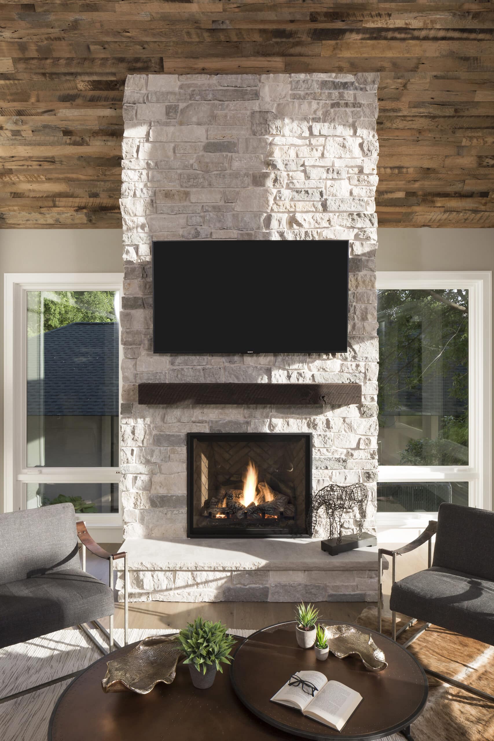 Stone fireplace with reclaimed timber mantel, hanging TV, and reclaimed wood paneling vaulted ceiling