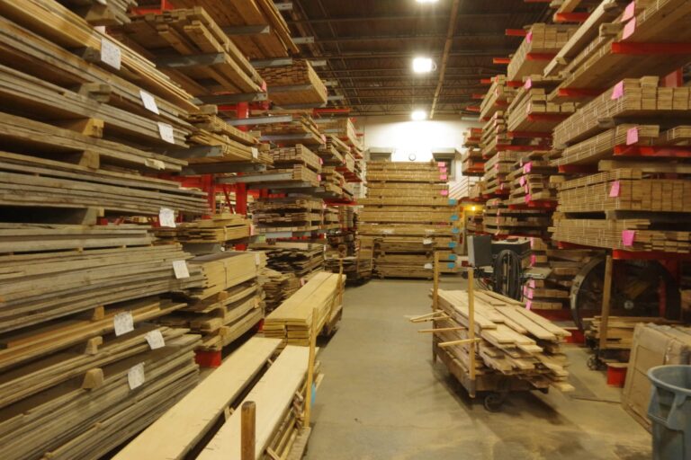Stacks of wooden boards, timber beams, and planks are organized on metal shelves in a warehouse with high ceilings and fluorescent lighting. A cart with wood pieces sits in the aisle. Pink labels mark many of the boards for interior design projects.