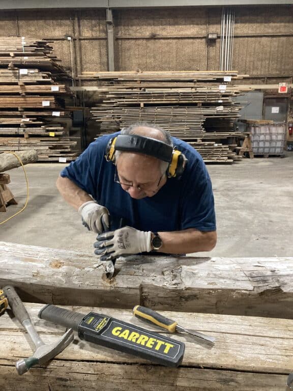 A man wearing gloves and ear protection carves wood with a chisel in a workshop, surrounded by stacks of lumber for wood mantels. Tools, including a hammer labeled GARRETT, are on the wooden beam in front of him.