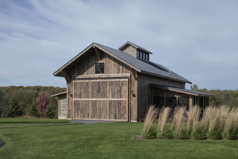 A rustic wooden barn with a metal roof stands on a lush green lawn, featuring reclaimed wood paneling, and is surrounded by trees and tall ornamental grasses under a partly cloudy sky.
