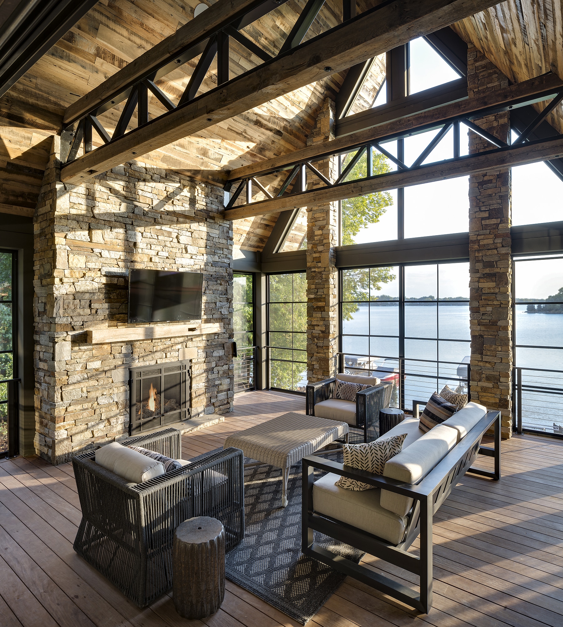 Lodge sitting room with glass wall view onto lake, vaulted ceilings with reclaimed wood paneling, steel trusses with wood box beam accents and large stone fireplace with timber mantel