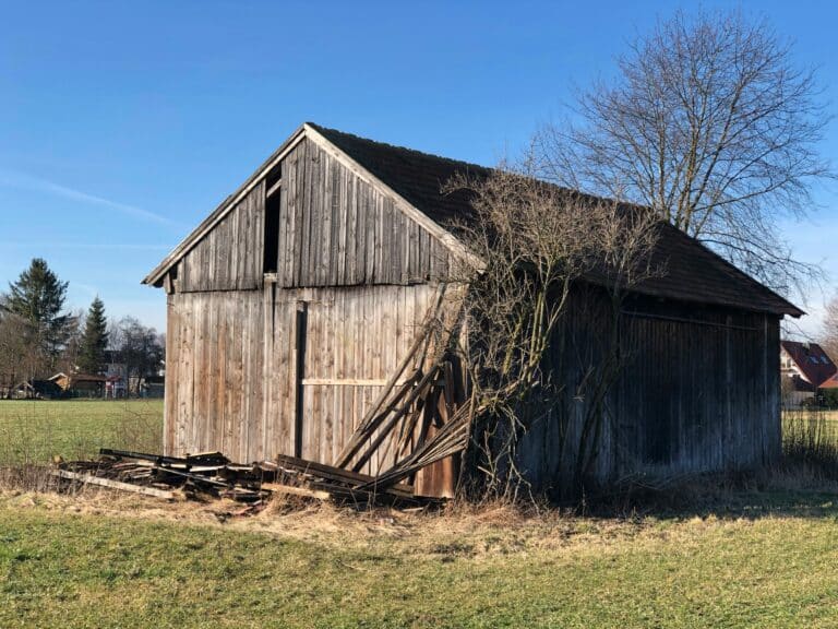 A weathered wooden barn with rustic wood paneling stands on a grassy field under a clear blue sky. Piles of old planks rest beside the barn, and leafless branches grow along its side. Trees and houses are visible in the background.