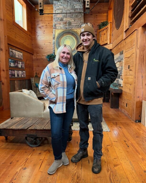 A woman and a man stand smiling inside a cozy, rustic wooden room with timber beams, a stone fireplace with a wood mantel, wooden floors, and art on the wall. The woman wears a plaid jacket and jeans; the man wears a beanie, black jacket, and brown pants.