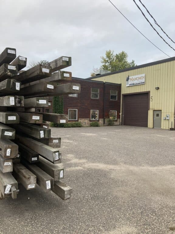A stack of large, labeled reclaimed wood beams sits in the parking lot outside a tan and brown industrial building with a sign that reads Manomin Resawn Timbers. Trees and a cloudy sky are visible in the background.