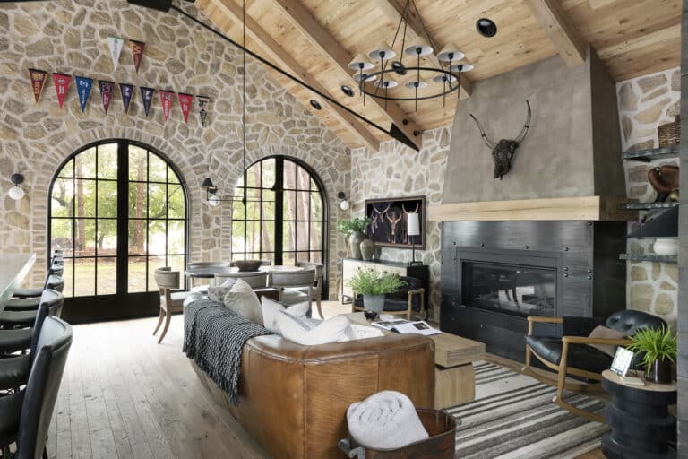 Warm home interior with stone walls, vaulted wood ceiling, and large arched windows letting in natural light; features a leather sofa, modern fireplace, and cozy interior design accents like blankets, plants, and rustic decor for true cozy vibes.