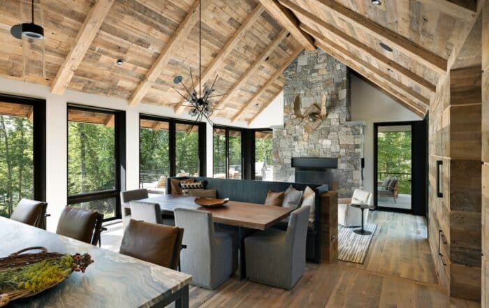 A cozy design with reclaimed wood throughout the space, featuring a large dining table and a comfortable seating nook by the stone fireplace. The room is filled with natural light from large windows, offering views of the surrounding trees. The high, wooden-beamed ceiling and modern decor create a warm, inviting atmosphere.