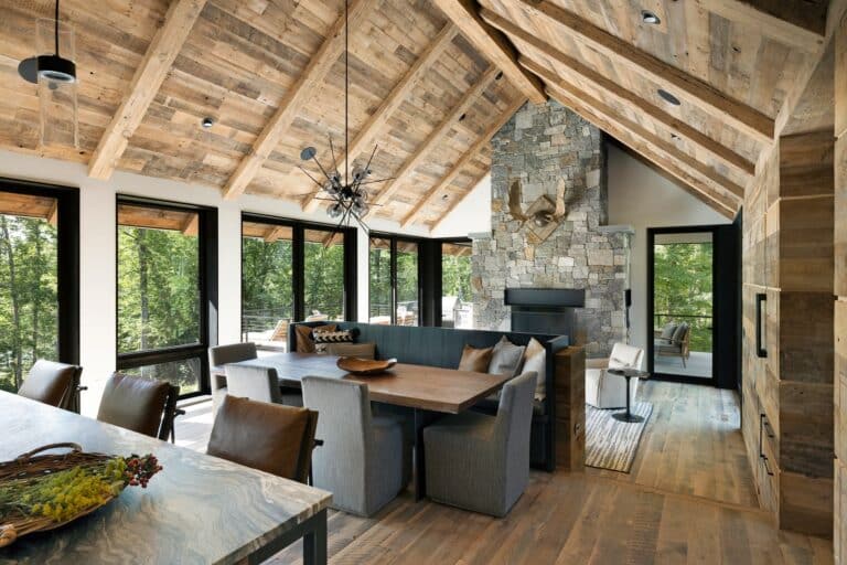 A cozy design with reclaimed wood throughout the space, featuring a large dining table and a comfortable seating nook by the stone fireplace. The room is filled with natural light from large windows, offering views of the surrounding trees. The high, wooden-beamed ceiling and modern decor create a warm, inviting atmosphere.