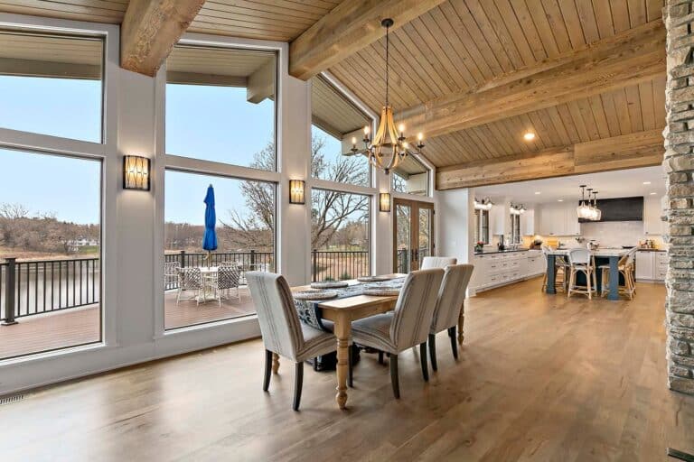 Dining room with vaulted ceiling and reclaimed wood beams, stone fireplace, and open kitchen design featuring large windows overlooking a deck, creating a warm home atmosphere.