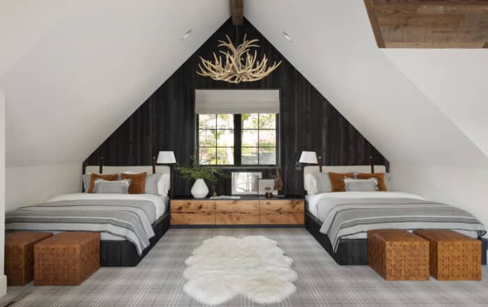 A cozy attic bedroom with two beds, plaid bedding, brown and white accents, a fur rug, antler chandelier, large window, and inviting wood paneling. The décor blends rustic and modern interior design styles.