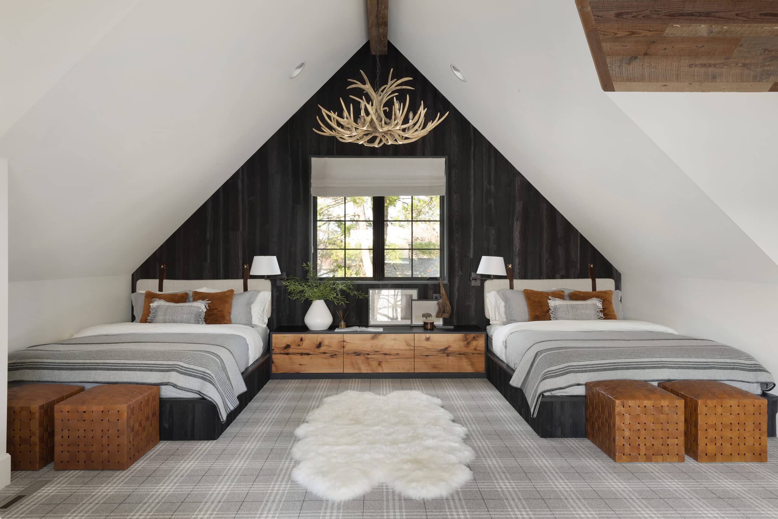 Wienzel Point Rd_066 A cozy attic bedroom with two beds, plaid bedding, brown and white accents, a fur rug, antler chandelier, large window, and inviting wood paneling. The décor blends rustic and modern interior design styles.