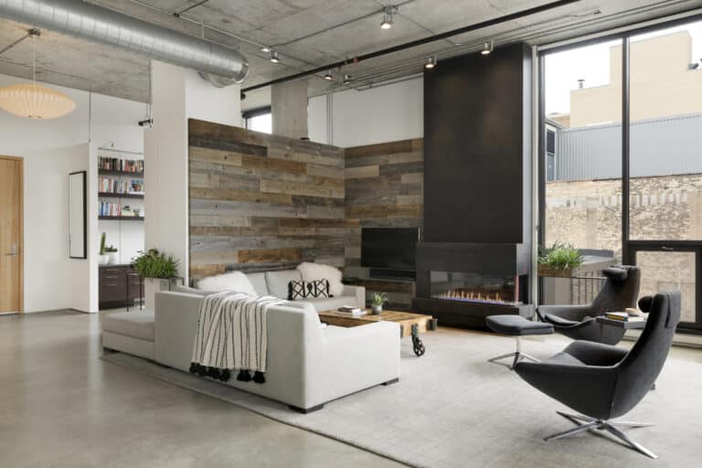 A modern loft living room featuring a reclaimed wood design accent wall behind a light-gray sectional sofa, offering stylish wall design ideas. The space includes an industrial concrete ceiling, a sleek black fireplace, large floor-to-ceiling windows, and contemporary lounge chairs, blending warm textures with minimalist decor.