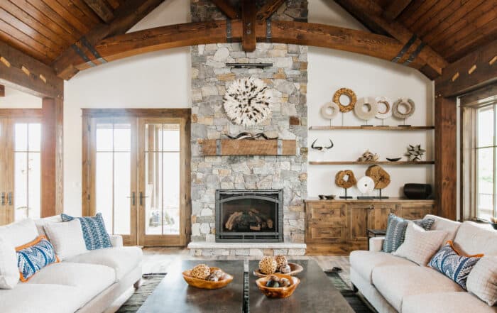 Rustic living room with a stone fireplace and wood fireplace mantel, wooden beams, neutral sofas, blue and white pillows, coffee tables with decor, and shelves displaying vases and woven art. Sunlight streams through large windows and glass doors.