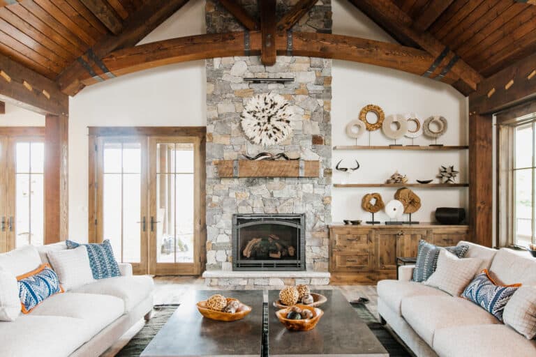 A cozy living room with a vaulted wood ceiling, featuring a stone fireplace as the centerpiece. The traditional interior design is enhanced by warm wood accents, including the wooden shelves displaying decorative pieces like horns and woven objects. The rich wood design is complemented by a modern sofa and coffee table arrangement, blending rustic elements with comfort. This space showcases classic wall design ideas and earthy tones for a welcoming atmosphere.
