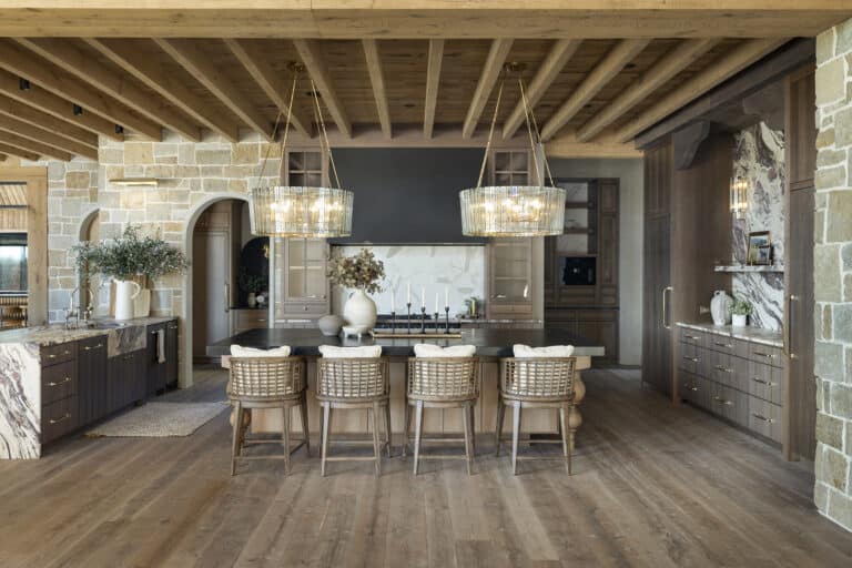 A spacious luxury kitchen featuring exposed wood ceiling beams, natural stone walls, a large island with woven chairs, and warm cabinetry, offering refined wood design inspiration with elegant lighting and earthy, rustic finishes
