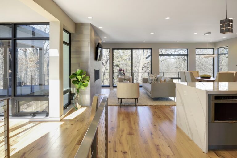 A modern living room with large windows allowing natural light to fill the space, featuring a cozy seating area with two chairs and a sofa. The warm wood flooring enhances the traditional interior design, while the contemporary wall design ideas are reflected in the sleek stone fireplace. The open space includes a kitchen area with a marble countertop, combining elements of wood design with minimalist accents.