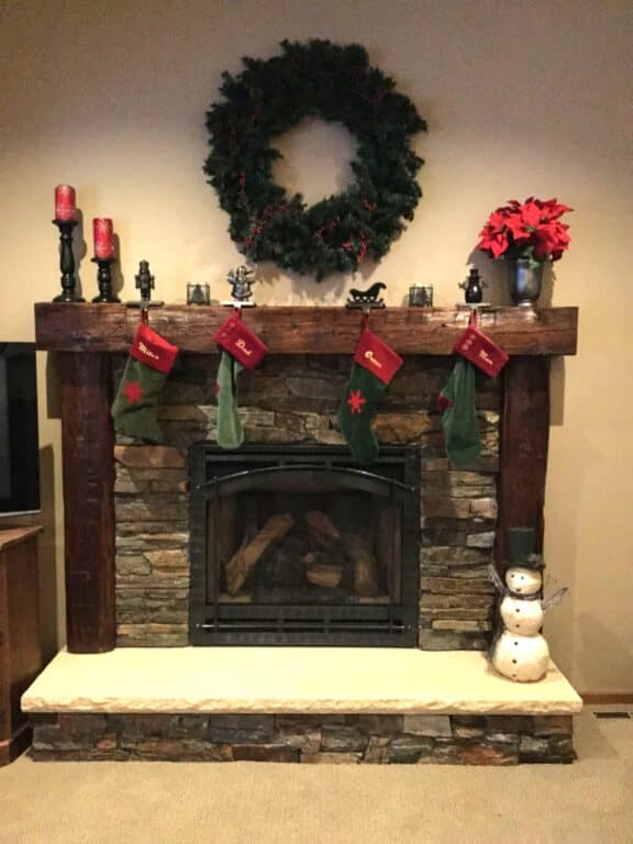 A stone fireplace with a reclaimed wood mantel decorated for the holidays, featuring green and red stockings, red candles, a large wreath, poinsettias, and rustic Christmas decorations including a small snowman figure