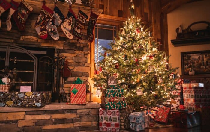 A warmly lit living room featuring a stone fireplace lined with colorful stockings and surrounded by glowing lights, wrapped gifts, and a beautifully decorated Christmas tree, creating a cozy scene of rustic Christmas decorations.