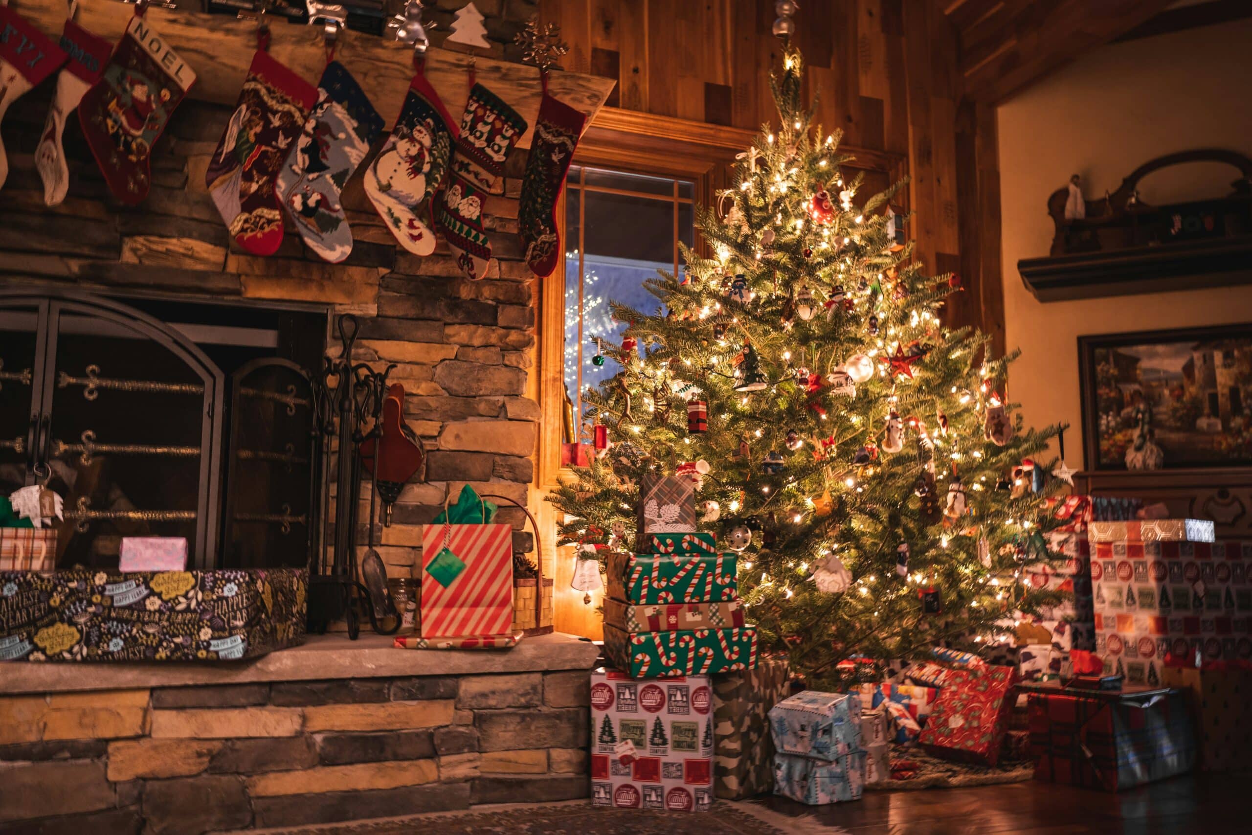 mike-cox-3UQybRpwpso-unsplash A warmly lit living room featuring a stone fireplace lined with colorful stockings and surrounded by glowing lights, wrapped gifts, and a beautifully decorated Christmas tree, creating a cozy scene of rustic Christmas decorations.
