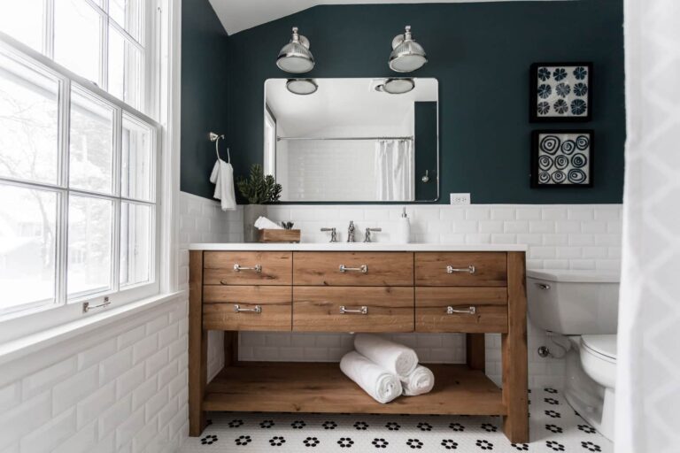 Modern bathroom with dark green upper walls, white subway tile, a large wood vanity with silver hardware, double sink, rolled white towels, large mirror, two industrial lights, custom cut wood shelving, and a window letting in natural light.