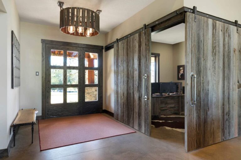 A modern rustic entryway features a glass-paneled custom cut wood double front door, a wooden bench, a red rug, and large sliding barn doors leading to an office with computers. A wooden chandelier hangs from the ceiling.