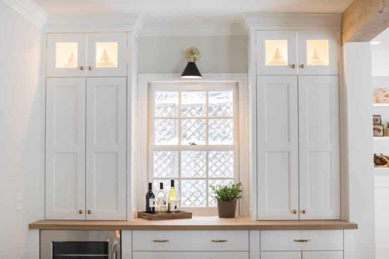 A kitchen with tall white cabinets flanking a window. A small potted plant and a tray with bottles are on the wooden countertop. The upper cabinets have glass panels displaying decorative items and are softly lit.