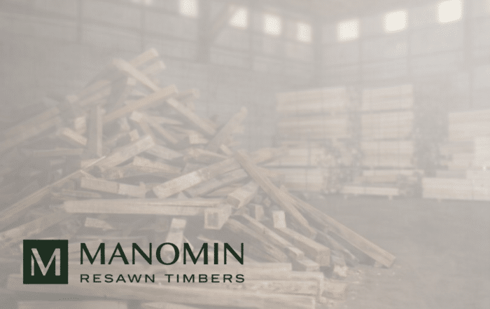 A pile of wooden beams inside a warehouse with stacks of timber in the background. In the foreground, the Manomin Resawn Timbers logo is displayed.