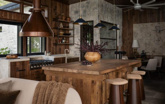 A rustic kitchen features a reclaimed wood wall, a large wooden island with stools, copper range hood, modern pendant lights, stone accents, open shelves, and a cozy seating area with a throw blanket.