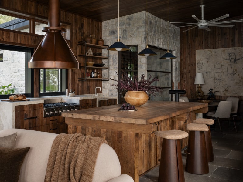 6LYnK7aw A rustic kitchen features a reclaimed wood wall, a large wooden island with stools, copper range hood, modern pendant lights, stone accents, open shelves, and a cozy seating area with a throw blanket.